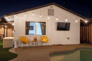 Una casa con piscina, dos sillas amarillas, una mesita con botellas y luces de cadena.
