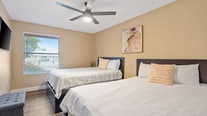 Une chambre avec deux lits, un ventilateur de plafond, une fenêtre avec des stores et un téléviseur fixé au mur.