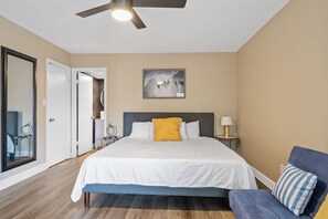 A bedroom with a bed, a nightstand, a lamp, a ceiling fan, a framed picture on the wall, and a door leading to another room.