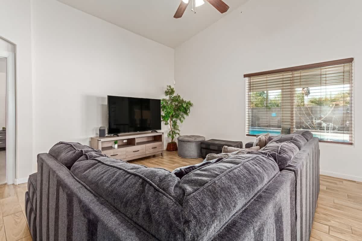 Una sala moderna con un sofá seccional gris grande, un televisor de pantalla plana sobre un mueble de madera, una planta en maceta y una ventana con persianas.
