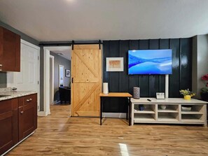 A room with a wooden floor, a sliding barn door, a television on a stand, and a small table with a vase. - Downtown Renovated Apt w/ Free Parking & BBQ (Phoenix)