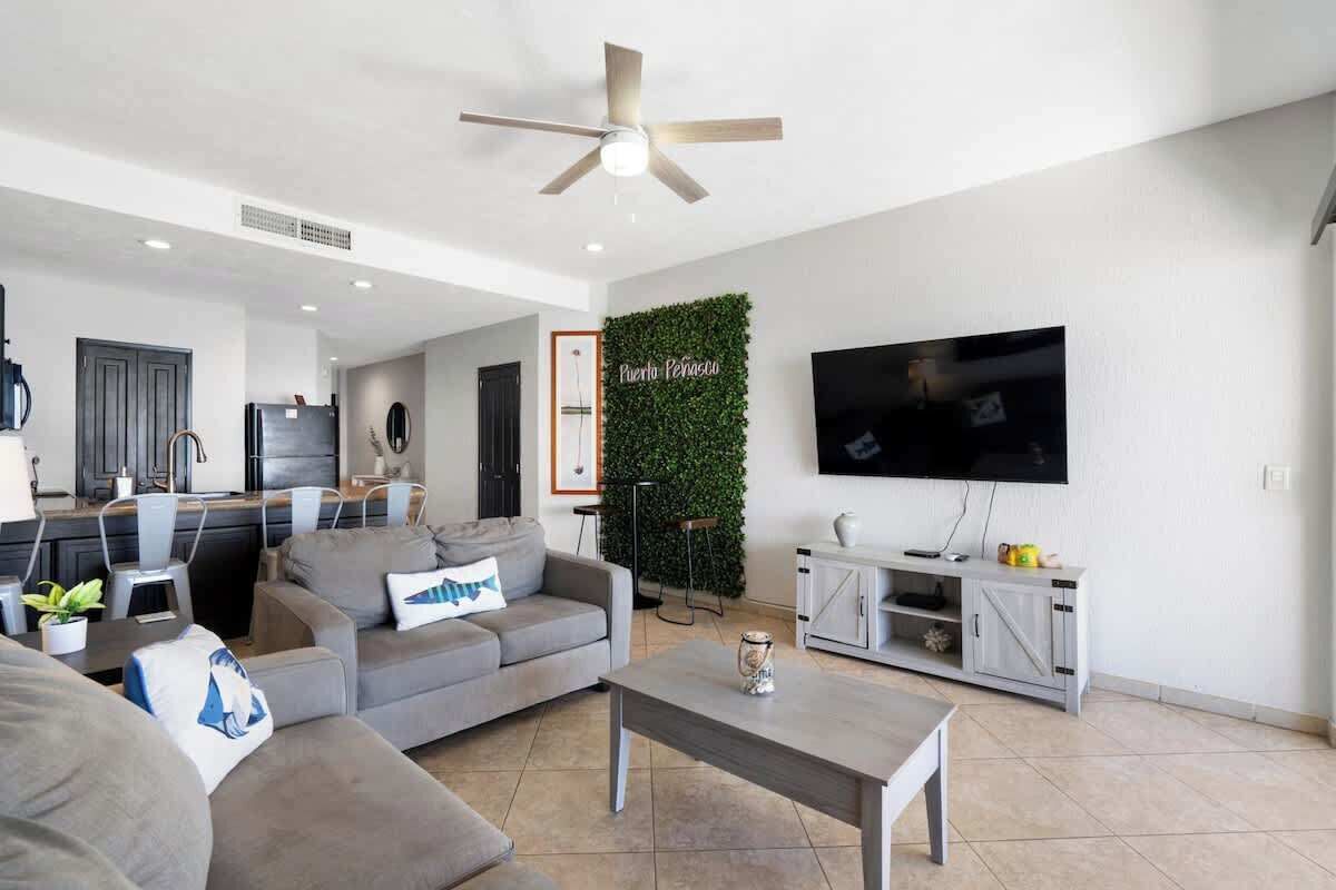 A modern living room with a grey sofa, a coffee table, a TV, and a kitchen area in the background.