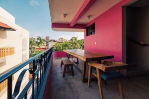 A balcony with a wooden table and benches, a railing, and a view of buildings and palm trees.