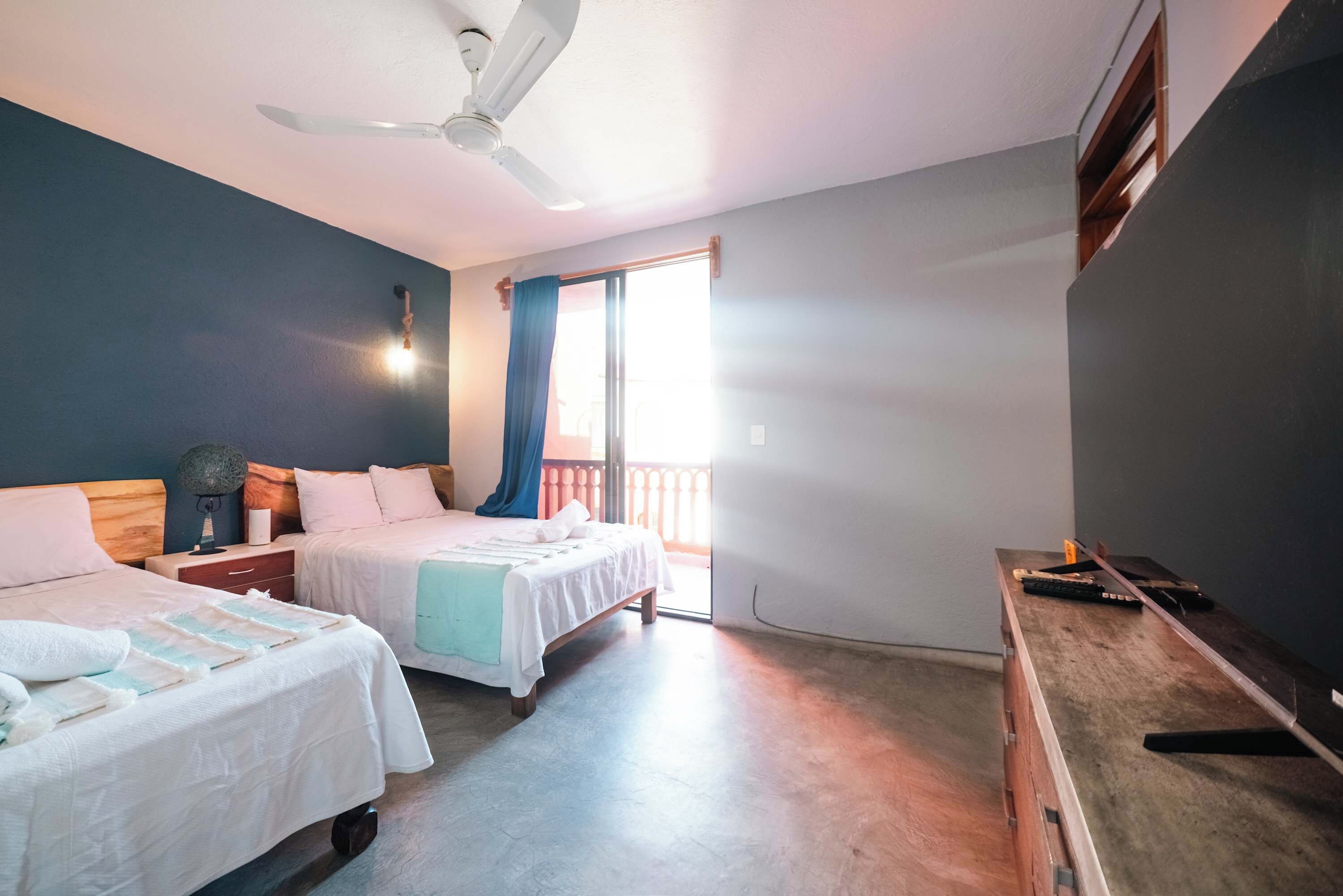 Un dormitorio con dos camas, un ventilador de techo, una mesita de noche, un aparador y una ventana con cortinas.