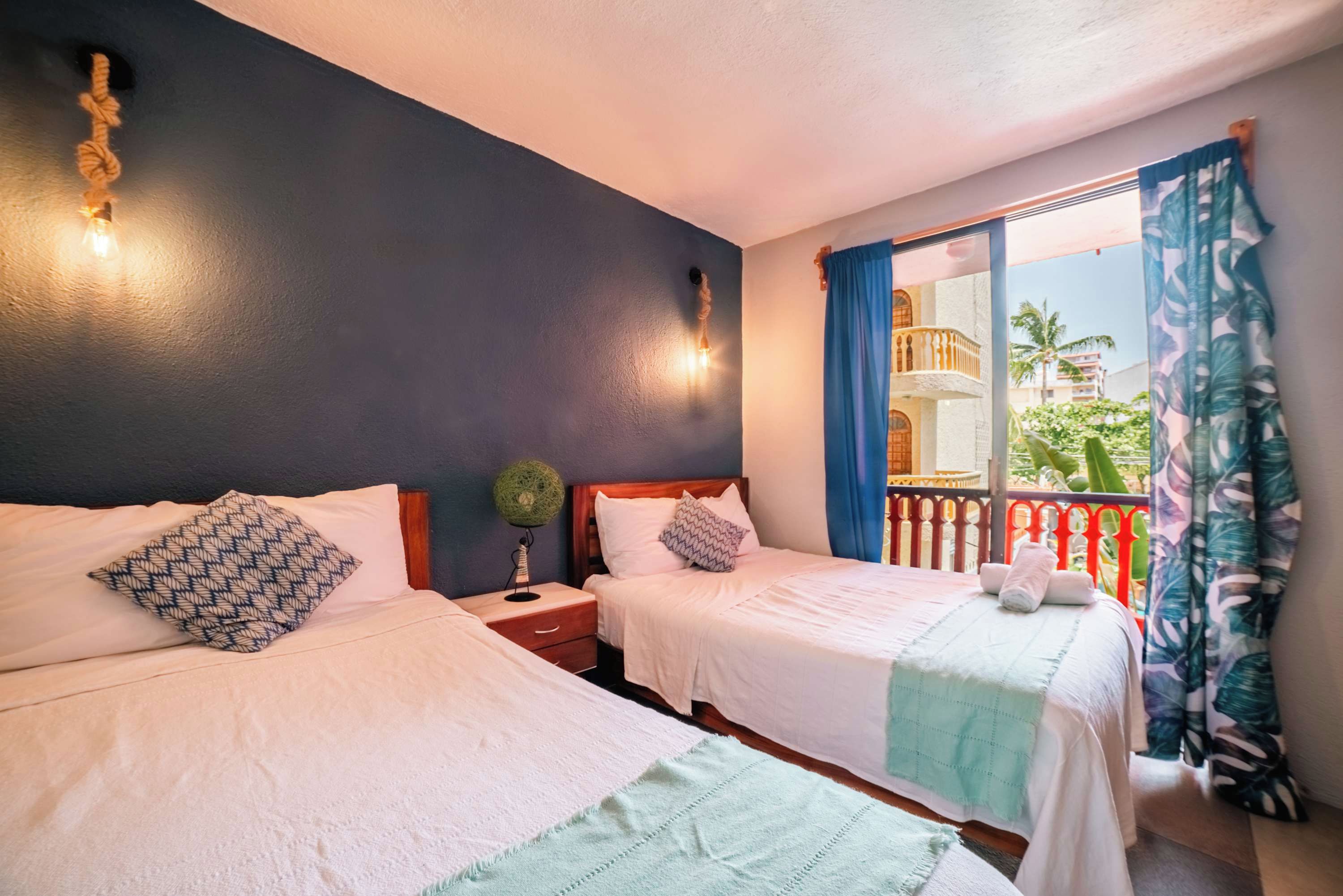 A hotel room with two beds, a nightstand with a lamp, a window with curtains, and a balcony with a railing.