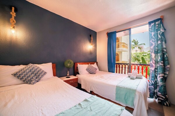 A hotel room with two beds, a nightstand with a lamp, a window with curtains, and a balcony with a railing.
