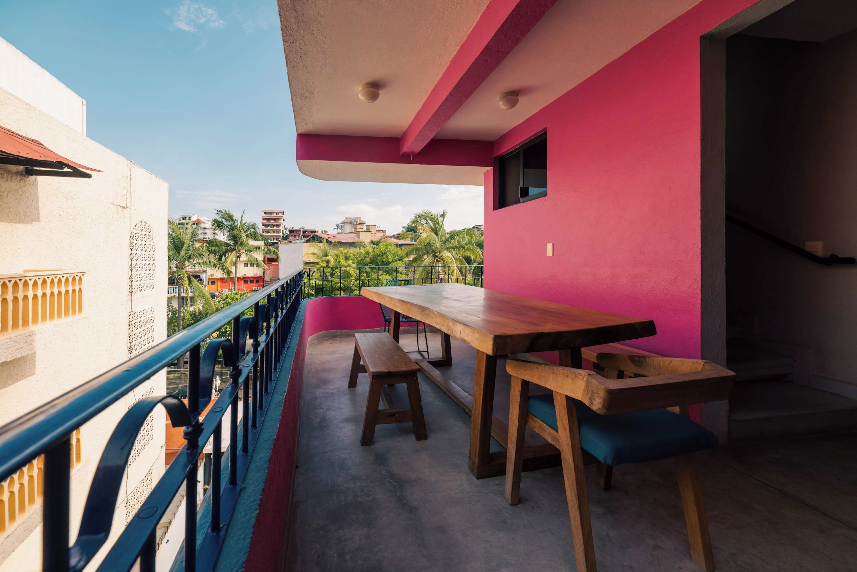 Un balcón con una mesa y bancos de madera, barandilla y vistas a edificios y palmeras.