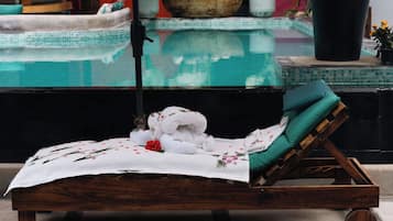 A wooden lounge chair with a green cushion, a white towel with floral patterns, and a red flower on a tiled floor.