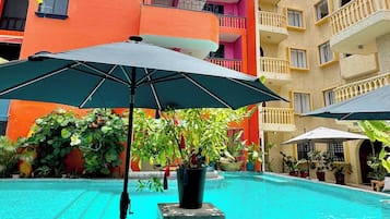 Zona junto a la piscina con una mesa y sillas, rodeada de plantas en macetas y un edificio de varios pisos con balcones.