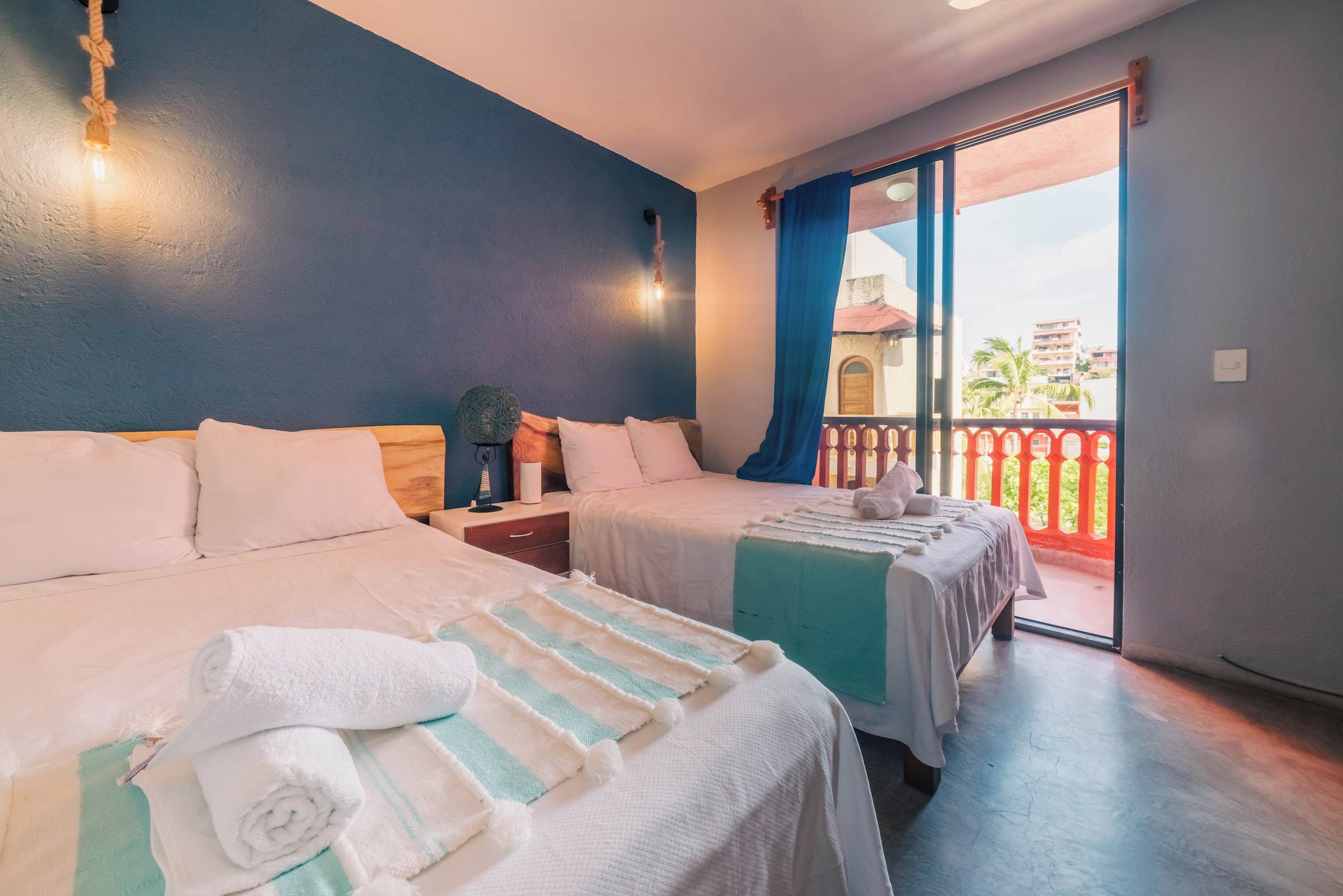 Habitación de hotel con dos camas, un balcón con barandilla roja y vistas a edificios.