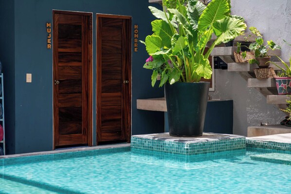 A pool area with a wooden door labeled 'Mujer' and 'Hombre,' a potted plant, and a tiled edge.