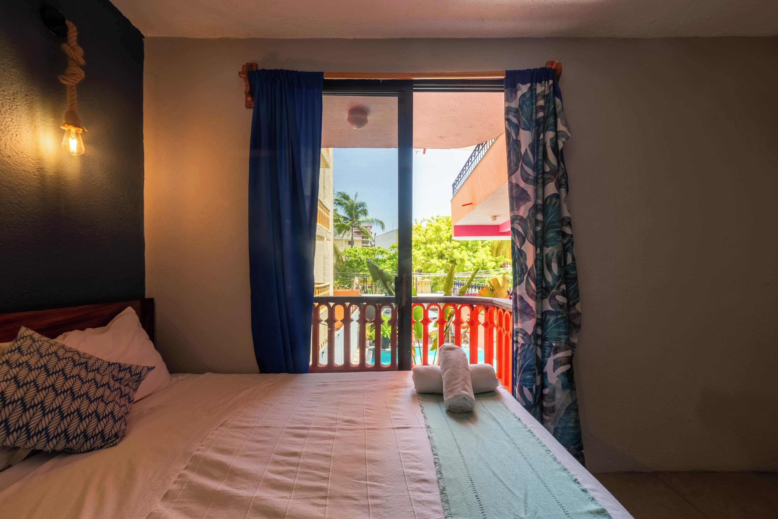 A hotel room with a bed, a window with curtains, a view of the outside, and a wall-mounted light fixture.