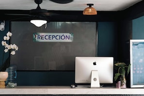 A reception desk with a computer, a water bottle, a vase with flowers, and a ceiling fan. - Zihua Mia Hotel Boutique 6 (Zihuatanejo)