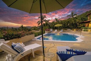 Un espace piscine agrémenté de chaises longues et de parasols, entouré d’une végétation luxuriante et offrant une vue imprenable sur le coucher de soleil.