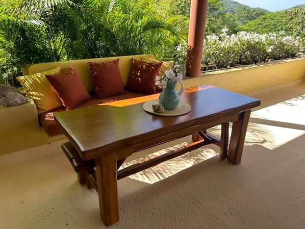 Une table en bois surmontée d’un vase de fleurs, un canapé avec des coussins et une vue sur la verdure et les collines.