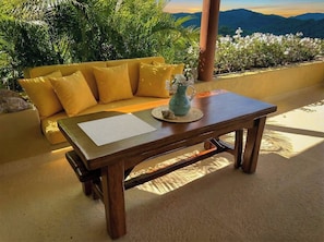 Ein Holztisch mit Unterlage und Dekoration, platziert auf einem Balkon mit gelbem Sofa und üppiger Bepflanzung im Hintergrund.