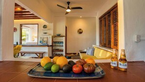 Une pièce comprenant une table à manger dressée avec des fruits et des bouteilles, un canapé, un ventilateur de plafond et une vue sur un jardin par la fenêtre.