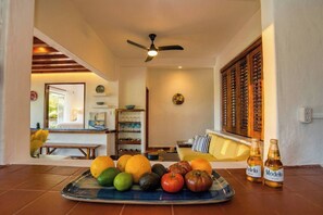 A room with a dining table set with fruits and bottles, a sofa, a ceiling fan, and a view of a garden through a window. - Villa Bougainvillea at Villas del Sol (Zihuatanejo)