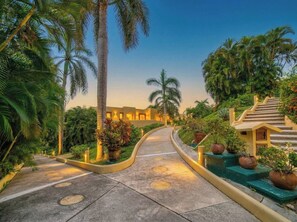A pathway leading to a building with a staircase on the right, surrounded by greenery and palm trees.