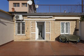 A residential building with a tiled patio, a covered outdoor seating area, and a visible door leading to an interior space.