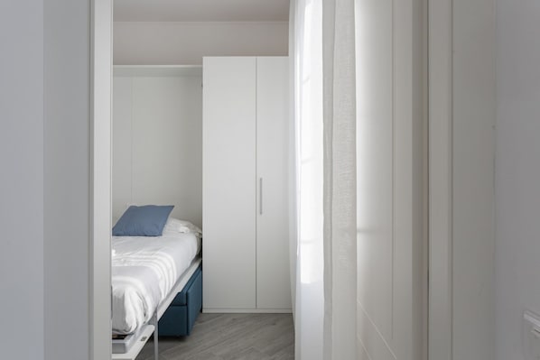 A small, narrow room with a bed, a wardrobe, and a window with sheer curtains.
