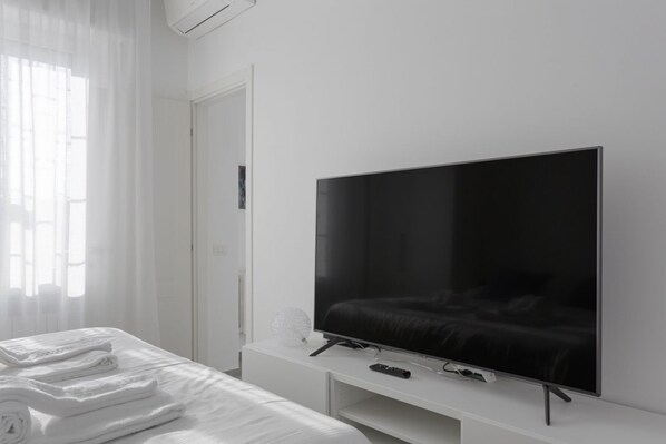 A modern hotel room with a large flat-screen TV on a white stand, a neatly made bed with white linens, and a window with sheer curtains.