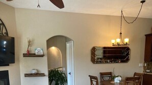 A spacious living area with a fireplace, a dining table with chairs, a kitchen area with a mirror, and a ceiling fan.