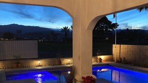 A pool area with a view of mountains at dusk, a patio with a table and chairs, and a decorative plant.