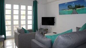 Un salon avec un canapé, un téléviseur à écran plat fixé au mur et un tableau représentant une scène de plage.