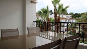 Un balcon avec une table et des chaises en verre, d’où l’on peut admirer des palmiers et un bâtiment blanc.