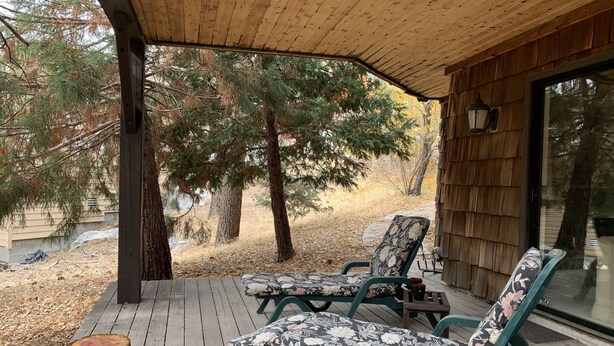 Une terrasse couverte avec deux chaises longues et une petite table, entourée d’arbres et d’un bâtiment en bois.