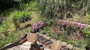 A garden with two wooden Adirondack chairs and a small table, surrounded by pebbles and various plants.