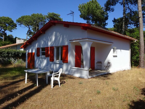 Une maison blanche aux volets rouges, un petit patio avec une table et des chaises, et un ciel d’un bleu limpide.