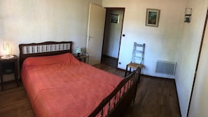 Un dormitorio con cama, mesita de noche, silla, radiador y puerta que da a otra habitación.