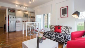 A modern kitchen with white cabinets, a dining area with white bar stools, a red sofa with a patterned throw, and a large window providing natural light. - Pacífico Apartamentos Ribera (Ushuaia)