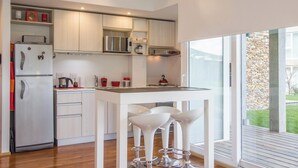 A modern kitchen with white cabinetry, a stainless steel refrigerator, a built-in microwave, and a coffee maker. There is a small island with bar stools and a window providing a view of the outdoors. - Pacífico Apartamentos Ribera (Ushuaia)
