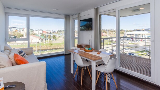 A modern living room with a large window, a dining area, and a flat-screen TV. - Pacífico Apartamentos Ribera (Ushuaia)