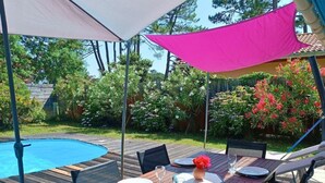 A wooden deck with a pool, surrounded by lush greenery and a pink shade sail. - pleasant villa of 120 qm for 8 people (BISCARROSSE)