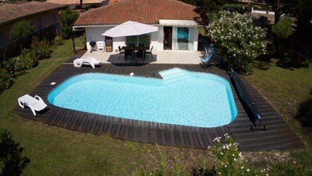 A rectangular swimming pool with a wooden deck, surrounded by a grassy area and a house with a tiled roof. - pleasant villa of 120 qm for 8 people (BISCARROSSE)