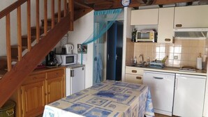 Una cocina con armarios de madera, un microondas, un tostador y un lavabo. Hay una mesa con mantel estampado. Se ve una escalera de madera al fondo.