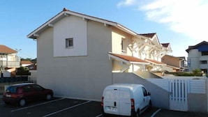 A two-story building with a white facade and a red-tiled roof. There is a white van parked in front, and a red car is visible in the background. - Beautiful duplex apartment of around 55m² for 4 people (BISCARROSSE)