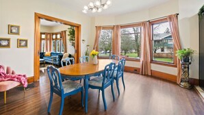 A dining area with a wooden table and blue chairs, a chandelier, and large windows with curtains. - Casa Van Gogh: a Historic N. Slope retreat (Tacoma)