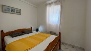 A bedroom with a wooden bed, a yellow blanket, a white pillow, a bedside table with a lamp, and a window with white curtains.