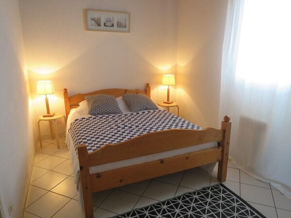 Un lit bien fait, recouvert d’une courtepointe à carreaux, avec deux lampes de chevet et un tableau encadré accroché au mur.
