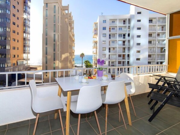 Un balcón con una mesa para dos, sillas blancas y vistas al mar y edificios.