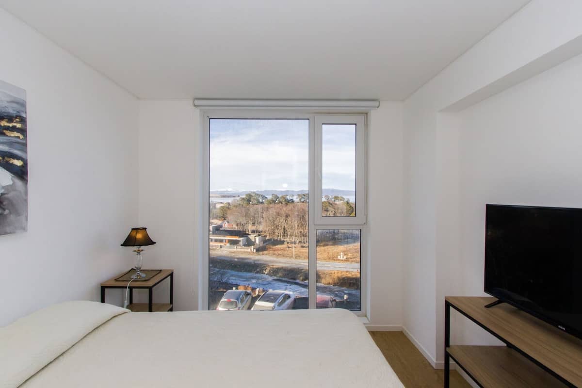 Un dormitorio con cama, televisor sobre un mueble de madera, lámpara en una mesita de noche y un ventanal con vista a un estacionamiento y edificios.