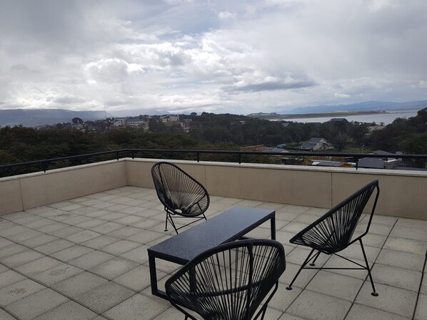 A balcony with two black wire mesh chairs and a rectangular table, overlooking a scenic view of a lake and distant mountains under a cloudy sky. - Pacífico Apartamentos Terrazas (Ushuaia)