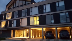 Een modern gebouw met twee verdiepingen, een plat dak, grote ramen en een overdekte parkeerplaats met twee auto's.