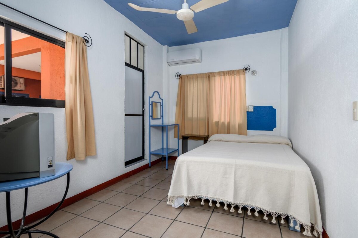 Habitación de hotel con una cama, un escritorio con computadora, un ventilador de techo y una ventana con cortinas.