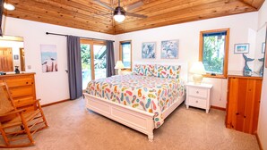Une chambre avec un plafond en bois, un lit avec une literie aux couleurs vives, un fauteuil à bascule, une table de chevet avec une lampe et une commode.